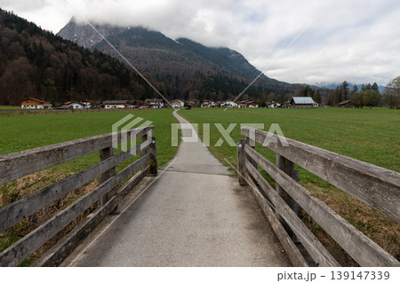 Rural path leading through green meadow toward alpine village with mountains and cloudy sky in Bavaria Germany 139147339