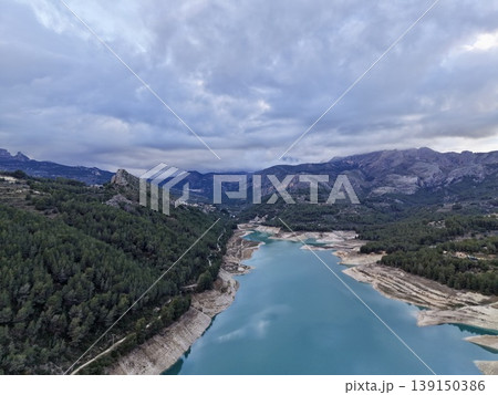 Calm mountain water amid dense forest shadows, Serene lake scene with rocky edges and overcast skies Calm mountain water amid dense forest shadows, Serene lake scene with rocky edges and overcast skies 139150386