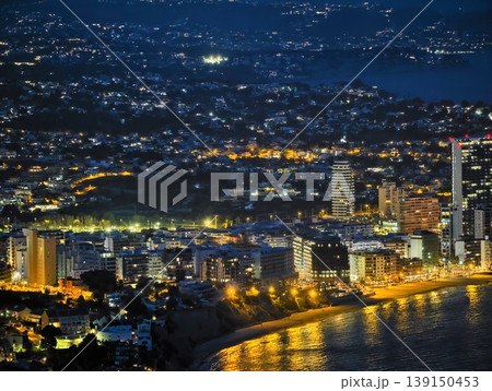 Modern buildings along shoreline. Vibrant city illumination reflecting on tranquil ocean at nighttime Modern buildings along shoreline. Vibrant city illumination reflecting on tranquil ocean at nighttime 139150453