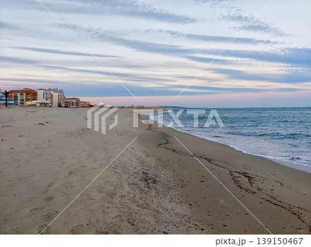 Gentle waves and sunset skies. Peaceful beach under pastel sky with serene ocean and distant structures Gentle waves and sunset skies. Peaceful beach under pastel sky with serene ocean and distant structures 139150467