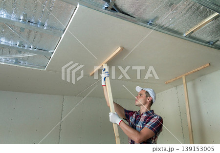 Young worker in protection work gloves fixing wooden holders for drywall suspended ceiling to metal frame insulated with shiny aluminum foil. DIY, do it yourself concept. 139153005
