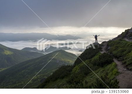 Wide mountain panorama. Small silhouette of tourist with backpack on rocky mountain slope with raised hands over valley covered with white puffy clouds. Beauty of nature, tourism and traveling concept 139153060