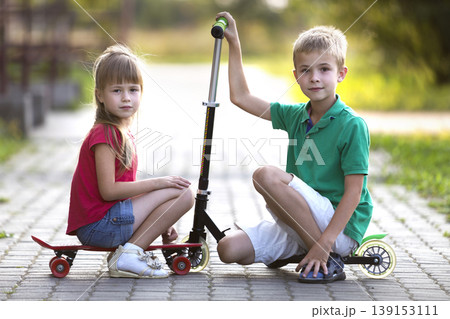 Two cute happy funny smiling young children, brother and sister, posing for camera, handsome boy with scooter and pretty long-haired girl sitting on skateboard on sunny colorful blurred background. 139153111