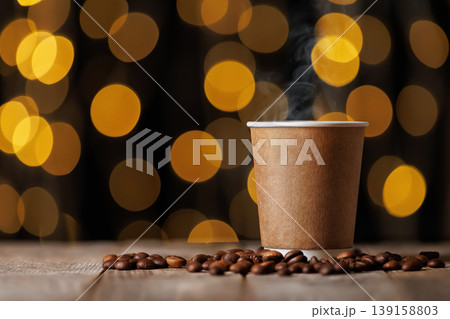 Steaming hot coffee drink in a blank paper cup, surrounded by roasted coffee beans on wooden table, with warm bokeh lights creating cozy atmosphere in the background Steaming hot coffee drink in a blank paper cup, surrounded by roasted coffee beans on wooden table, with warm bokeh lights creating cozy atmosphere in the background 139158803