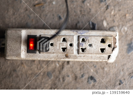 Close up of old power strip with glowing switch and worn sockets on dusty floor electrical safety risk and aging equipment in workshop environment Close up of old power strip with glowing switch and worn sockets on dusty floor electrical safety risk and aging equipment in workshop environment 139159640