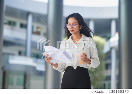 Stylish office employee holding documents, pen, and iced coffee in an urban business courtyard, presenting organized workflow, focus, and everyday professional efficiency outdoors daily. Stylish office employee holding documents, pen, and iced coffee in an urban business courtyard, presenting organized workflow, focus, and everyday professional efficiency outdoors daily. 139160379