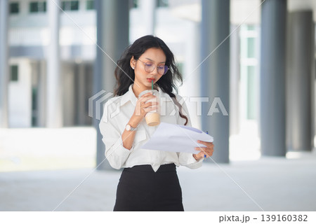 Young office professional drinking iced coffee and reading documents outdoors, showing focused work, daily routine, and modern business lifestyle in a bright corporate architectural setting Young office professional drinking iced coffee and reading documents outdoors, showing focused work, daily routine, and modern business lifestyle in a bright corporate architectural setting 139160382