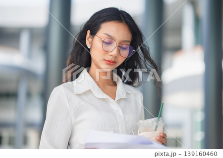 Stylish businesswoman in glasses reviewing papers and holding iced coffee in front of a modern office, illustrating thoughtful analysis, workplace focus, and professional urban lifestyle. Stylish businesswoman in glasses reviewing papers and holding iced coffee in front of a modern office, illustrating thoughtful analysis, workplace focus, and professional urban lifestyle. 139160460
