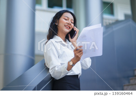 Smiling businesswoman talking on smartphone while reviewing paperwork outside a modern office building, capturing professional communication, mobility, and confident corporate workflow in daylight. 139160464