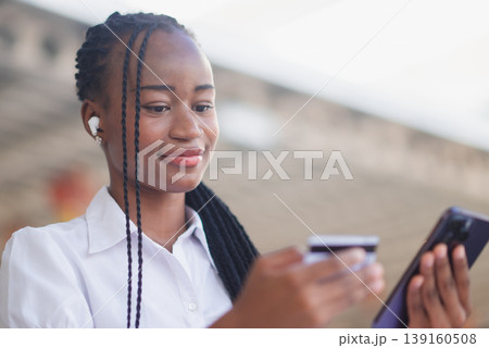 Young Black business woman using smartphone and credit card at outdoor cafe table with tablet nearby. Online payment, mobile banking, digital commerce and small business lifestyle concept. Young Black business woman using smartphone and credit card at outdoor cafe table with tablet nearby. Online payment, mobile banking, digital commerce and small business lifestyle concept. 139160508