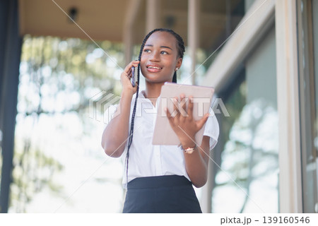 Young Black business woman on smartphone call and using tablet outdoors in front of modern office. Client communication, remote coordination, digital planning and mobile business workflow concept. Young Black business woman on smartphone call and using tablet outdoors in front of modern office. Client communication, remote coordination, digital planning and mobile business workflow concept. 139160546