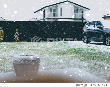 Snowfall seen through a window onto a suburban yard, a modern house and parked car. Winter weather, cold season, real estate and housing theme. 139161471