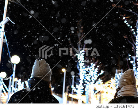 A candid, over-the-shoulder shot of people in winter clothing admiring a brightly lit outdoor urban scene at night. Snowfall and Christmas lights.  139161473