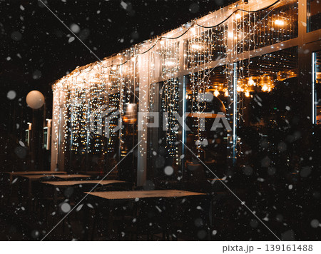 Cozy winter evening ambiance at an outdoor cafe terrace during snowfall. The scene is illuminated by glowing string fairy lights. Cafe patio, terrace. Cozy winter evening ambiance at an outdoor cafe terrace during snowfall. The scene is illuminated by glowing string fairy lights. Cafe patio, terrace. 139161488