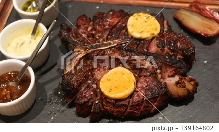 close up of a Grilled steak served on slate plate with melting herb butter, coarse salt, roasted shallot, and assorted sauces in ramekins of cream, pestle sauce and tomato sauce 139164802