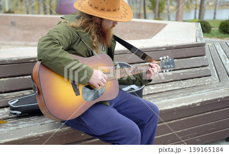 A red-haired, bearded guy in a cowboy hat is playing guitar in the park. A street musician. The guitarist gives guitar lessons outdoors. The concept of inspiration 139165184