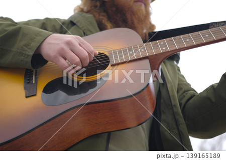 View of the guitarist from below. A man with a red beard plays the guitar, the focus is on the hand and the strings. The musician gives guitar lessons outdoors. The bearded man 139165189