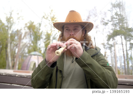 A red-haired man with curly hair and a beard is playing a flute. A musician in a cowboy hat plays a pipe in the park. The image is made in a rustic and peaceful style 139165193