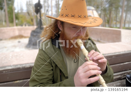 A thoughtful bearded guy in a cowboy hat plays a pipe in the park sitting on a bench. The musician plays the flute. The flutist has caught inspiration and is composing a new melody A thoughtful bearded guy in a cowboy hat plays a pipe in the park sitting on a bench. The musician plays the flute. The flutist has caught inspiration and is composing a new melody 139165194