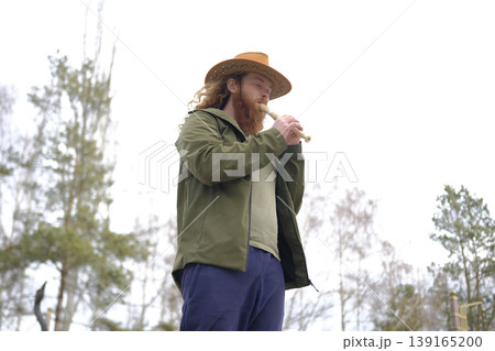 A handsome red-bearded man in a cowboy hat is playing the flute in the park. The piper plays the pipe, bottom view. Portrait of a free musician dressed in a green jacket A handsome red-bearded man in a cowboy hat is playing the flute in the park. The piper plays the pipe, bottom view. Portrait of a free musician dressed in a green jacket 139165200