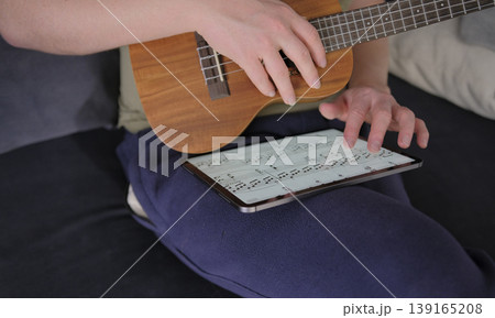 A man with a ukulele in his hands is looking through the notes of a new melody on a tablet. A guitarist learns to play the ukulele. The musician is sitting on the couch A man with a ukulele in his hands is looking through the notes of a new melody on a tablet. A guitarist learns to play the ukulele. The musician is sitting on the couch 139165208