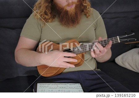 Ukulele lessons. A man with curly red hair and a beard is playing the ukulele sitting on the couch. The guitarist learns a new melody from the notes on the tablet 139165210