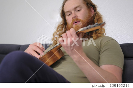 A man with a red curly beard and wavy red hair is playing the ukulele in his room. He is sitting on a blue sofa and carefully playing chords. The guitarist is learning a new tune A man with a red curly beard and wavy red hair is playing the ukulele in his room. He is sitting on a blue sofa and carefully playing chords. The guitarist is learning a new tune 139165212