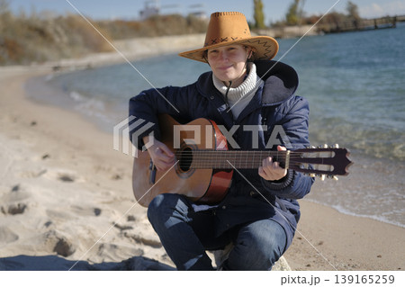 A young woman is sitting on the beach with a guitar in her hands. She's wearing a brown cowboy hat and a blue jacket. The guitarist plays on the shore of a blue lake 139165259