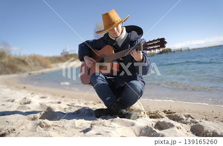Guitar lessons. A young mysterious woman in a cowboy hat is playing guitar on the beach by the ocean. The scene is calm and relaxing 139165260
