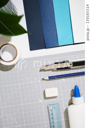 Craft supplies flat lay on cutting mat: utility knife, pencil, eraser, glue, tape, ruler, and blue cardstock sheets. Top view, white background. Craft supplies flat lay on cutting mat: utility knife, pencil, eraser, glue, tape, ruler, and blue cardstock sheets. Top view, white background. 139165314