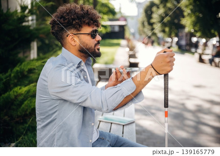 Smartphone in hand, talking. Blind man with white cane and sunglasses is in the city Smartphone in hand, talking. Blind man with white cane and sunglasses is in the city 139165779