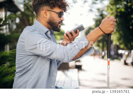 Smartphone in hand, talking. Blind man with white cane and sunglasses is in the city Smartphone in hand, talking. Blind man with white cane and sunglasses is in the city 139165781