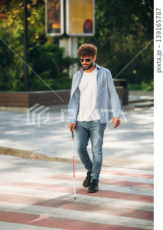 Wearing casual clothes, having a walk. Blind man with white cane and sunglasses is in the city 139165787