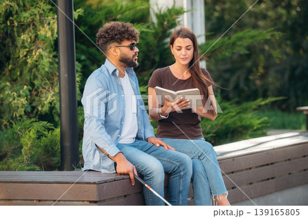 Interesting information. Woman is with blind man, sitting outdoors and reading book 139165805