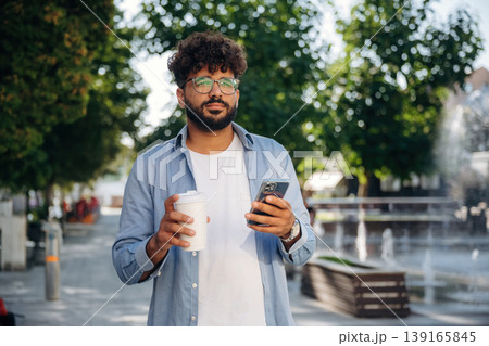 Cup of drink and smartphone are in hands. Handsome man is outdoors in the city 139165845