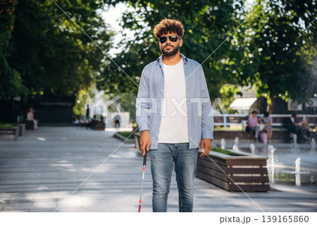 Front view, walking. Blind man with white cane and sunglasses is in the city 139165860