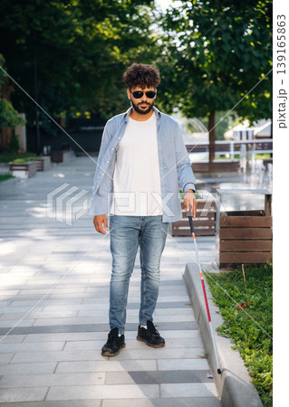 Front view, walking. Blind man with white cane and sunglasses is in the city Front view, walking. Blind man with white cane and sunglasses is in the city 139165863