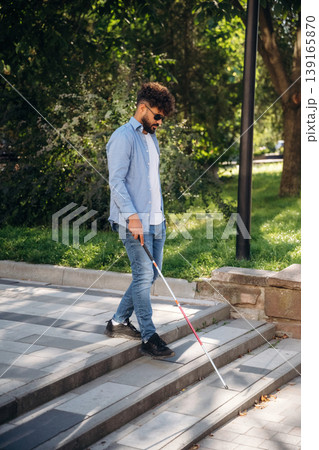 Having a walk, using cane stick. Blind man with white cane and sunglasses is in the city 139165870