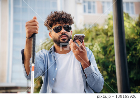 Communication, holding smartphone. Blind man with white cane and sunglasses is in the city Communication, holding smartphone. Blind man with white cane and sunglasses is in the city 139165926
