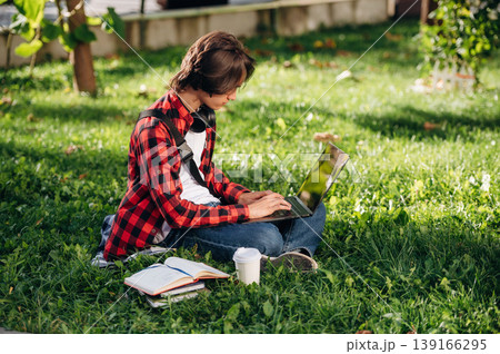 Laptop is on legs. Male student is in the public park outdoors Laptop is on legs. Male student is in the public park outdoors 139166295