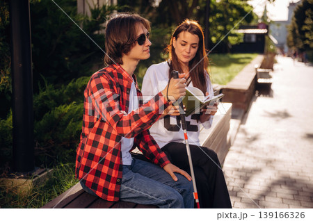 Woman is reading the book for blind guy, sitting outdoors 139166326