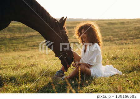 Side view, sitting on the ground. Woman in white dress is with horse on the countryside field 139166685