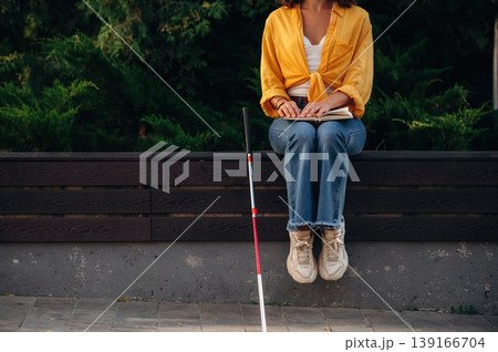 Blind woman with white cane is sitting and reading the book using Braille language and technique Blind woman with white cane is sitting and reading the book using Braille language and technique 139166704