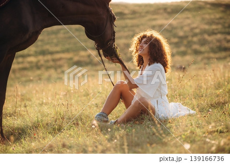 Side view, sitting on the ground. Woman in white dress is with horse on the countryside field 139166736
