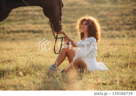 Side view, sitting on the ground. Woman in white dress is with horse on the countryside field Side view, sitting on the ground. Woman in white dress is with horse on the countryside field 139166739