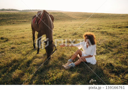 Resting, sitting on the ground. Woman in white dress is with horse on the countryside field Resting, sitting on the ground. Woman in white dress is with horse on the countryside field 139166761