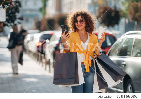 Cup of drink and smartphone in hands. Happy woman is with shopping bags in town Cup of drink and smartphone in hands. Happy woman is with shopping bags in town 139166986