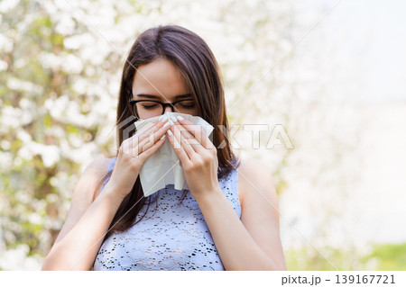 Sick young girl against blooming trees in spring. Outdoor shot of irritated female with seasonal allergy, uses white tissue, poses in light blouse with napkin, has rhinitis and sneezing Sick young girl against blooming trees in spring. Outdoor shot of irritated female with seasonal allergy, uses white tissue, poses in light blouse with napkin, has rhinitis and sneezing 139167721