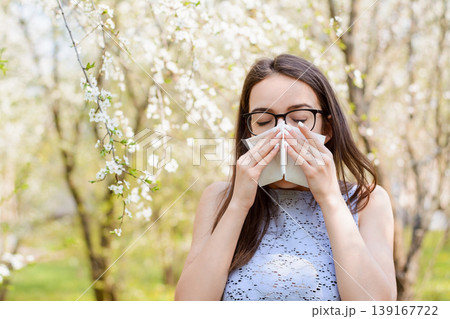 Young female with long natural hair in light blouse sneezing because of bloom pollen from trees in spring. Concept of allergy on pollen Young female with long natural hair in light blouse sneezing because of bloom pollen from trees in spring. Concept of allergy on pollen 139167722