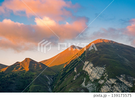 Mountain peaks bathed in warm golden light under a sky with soft orange clouds at an early morning or late evening Mountain peaks bathed in warm golden light under a sky with soft orange clouds at an early morning or late evening 139173575